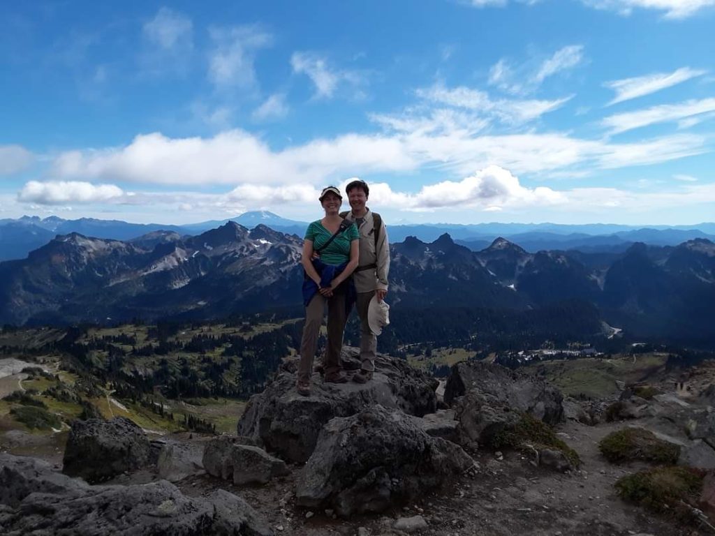 two people standing on high rock in center of image. background is long line of rocky mountain tops and blue sky with clouds.